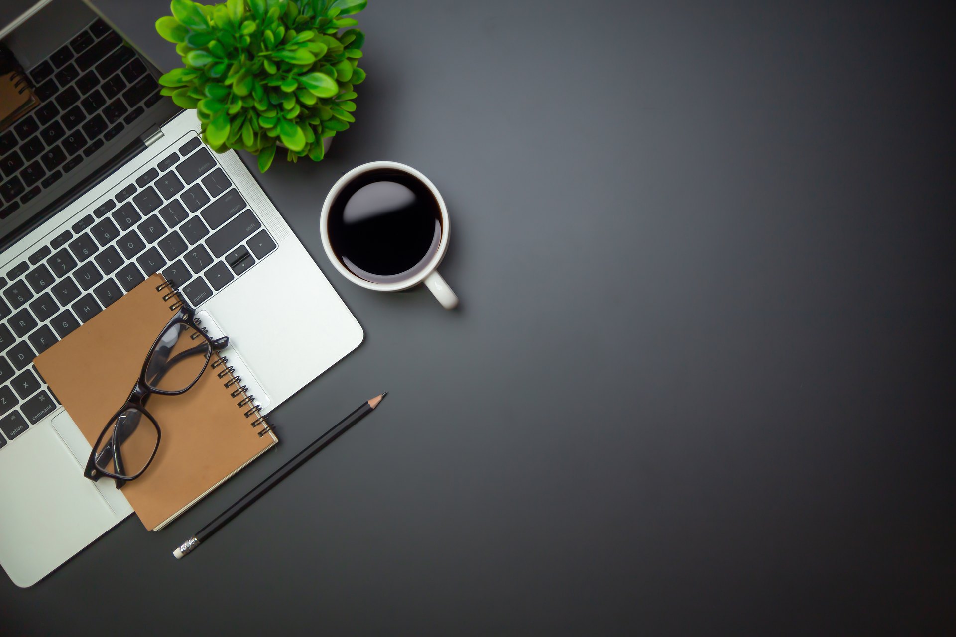 Dark desk with laptop, notebook, coffee and plant